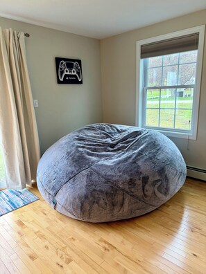 A room with a large bean bag chair, a window with blinds, a wall-mounted game controller art piece, and a wooden floor. - Charming Home with Private Neighborhood Beach (Naples)