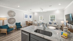 Eine moderne Küche mit Granit Arbeitsplatte und Edelstahlspüle, ein Essbereich mit Tisch und Stühlen sowie ein Wohnzimmer mit Sofa und Fernseher.
