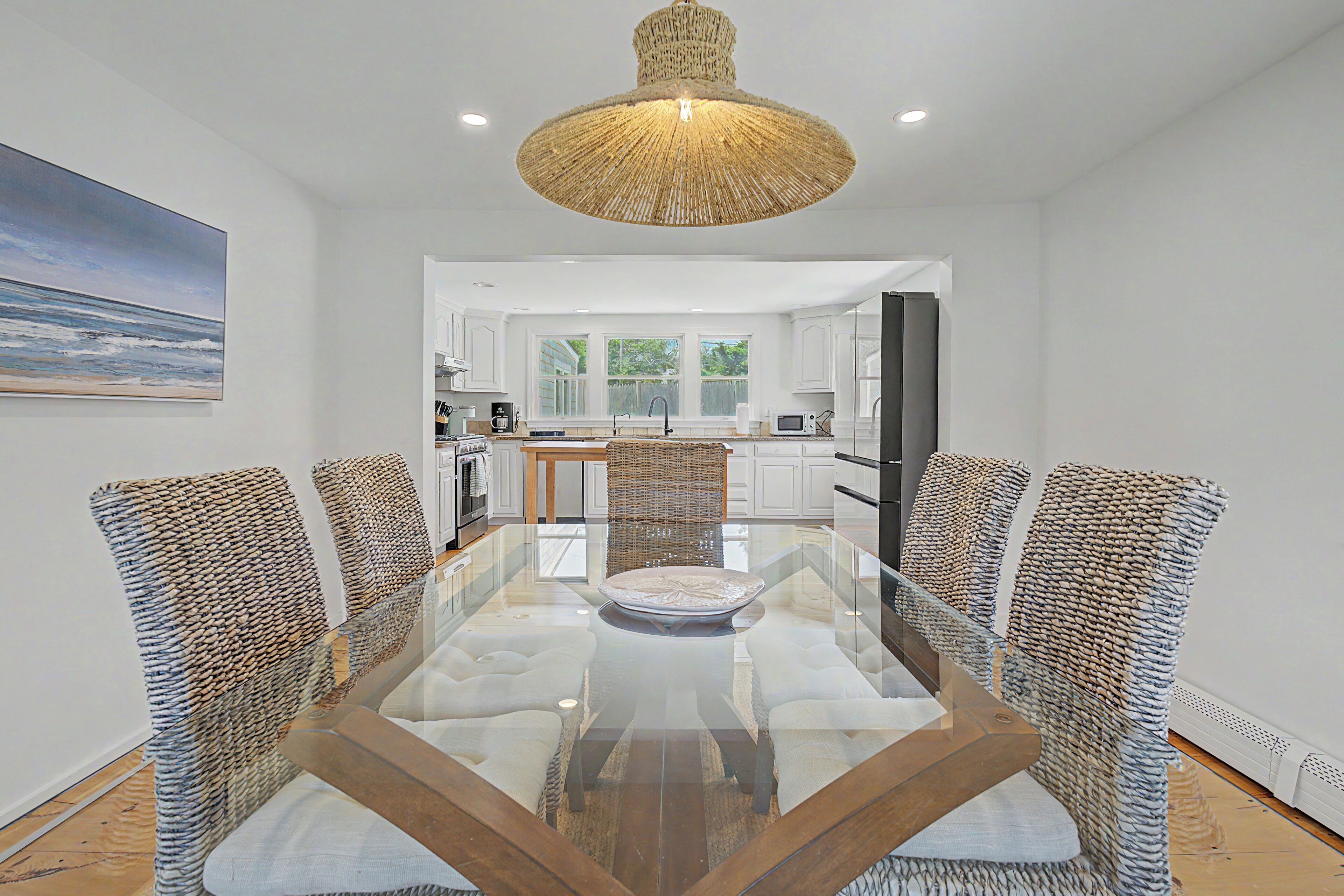 A dining area with a glass-top table surrounded by wicker chairs, a woven ceiling light fixture, and a view into a kitchen with white cabinets.