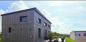 Un bâtiment moderne en bois, doté d’un toit plat, de nombreuses fenêtres et d’un espace extérieur pavé agrémenté de plantes en pot.