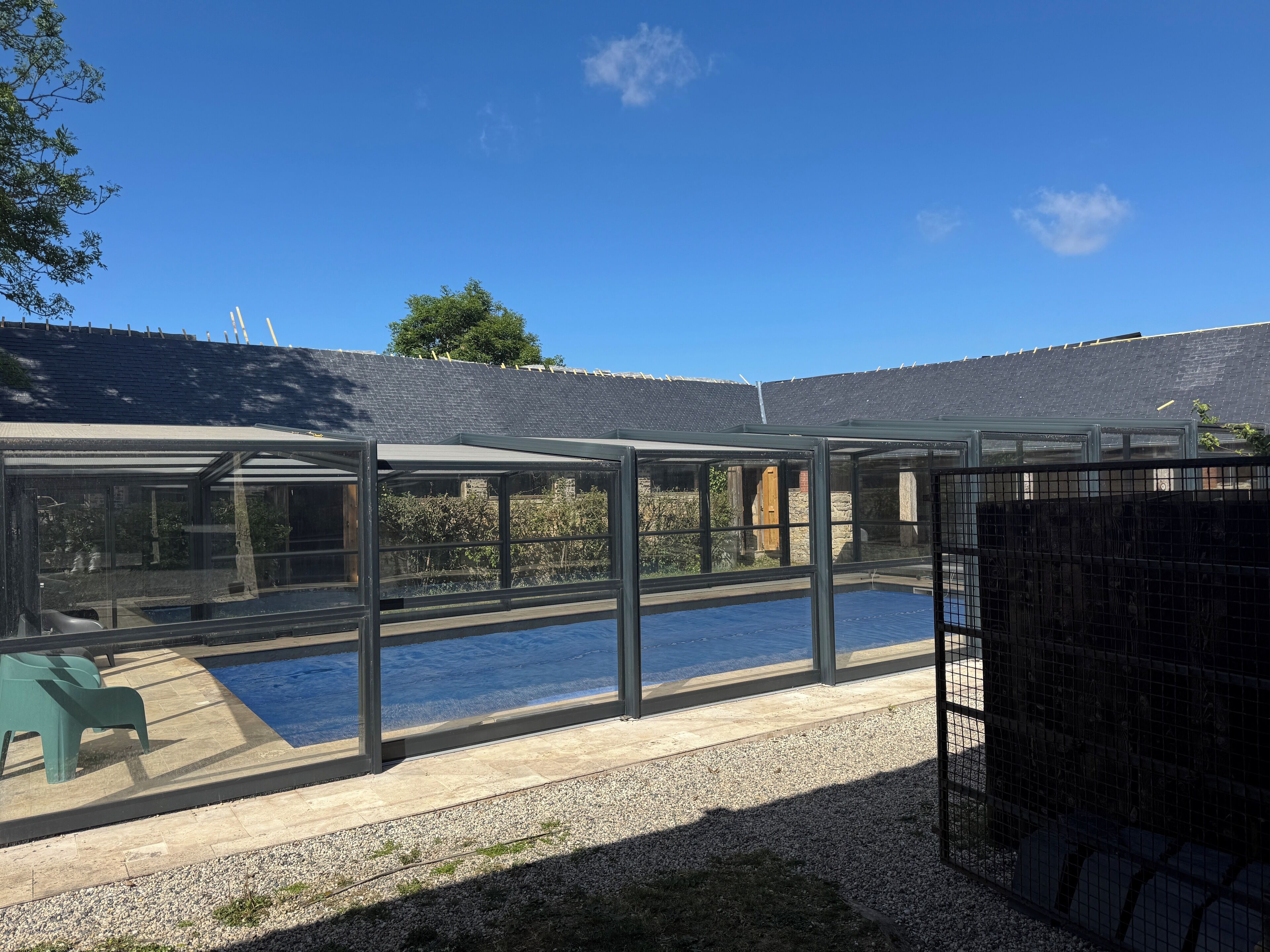 A covered outdoor swimming pool with a glass enclosure, surrounded by a gravel area and a stone wall.