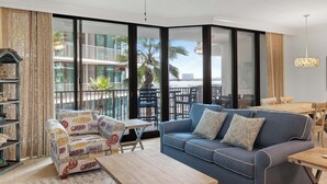 Un salon moderne avec un canapé bleu, un fauteuil à motifs et une table basse en bois. De grandes fenêtres offrent une vue sur un balcon aménagé avec du mobilier d’extérieur.