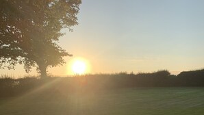 A circular stone fire pit with two benches, surrounded by a grassy field, with the sun setting in the background.