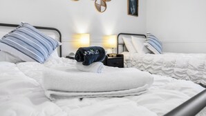 Een hotelkamer met twee bedden, witte beddengoed, een klein nachtkastje met een lamp, en wanddecoratie inclusief een lijst met een plant en een wanddecoratie van riet.