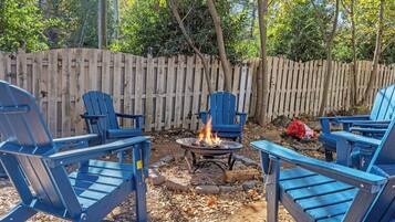 Un patio trasero con cerco de madera, una fogata y sillas Adirondack azules.