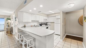 A modern kitchen with white cabinetry, a central island with bar stools, and a view of the ocean. - Coastal 2BR - pool, beach access & daily tickets (Panama City Beach)