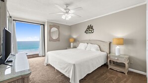 A bedroom with a large bed, a television, a desk, a ceiling fan, a window with a view of the ocean, and a wall-mounted ship decoration. - Coastal 2BR - pool, beach access & daily tickets (Panama City Beach)