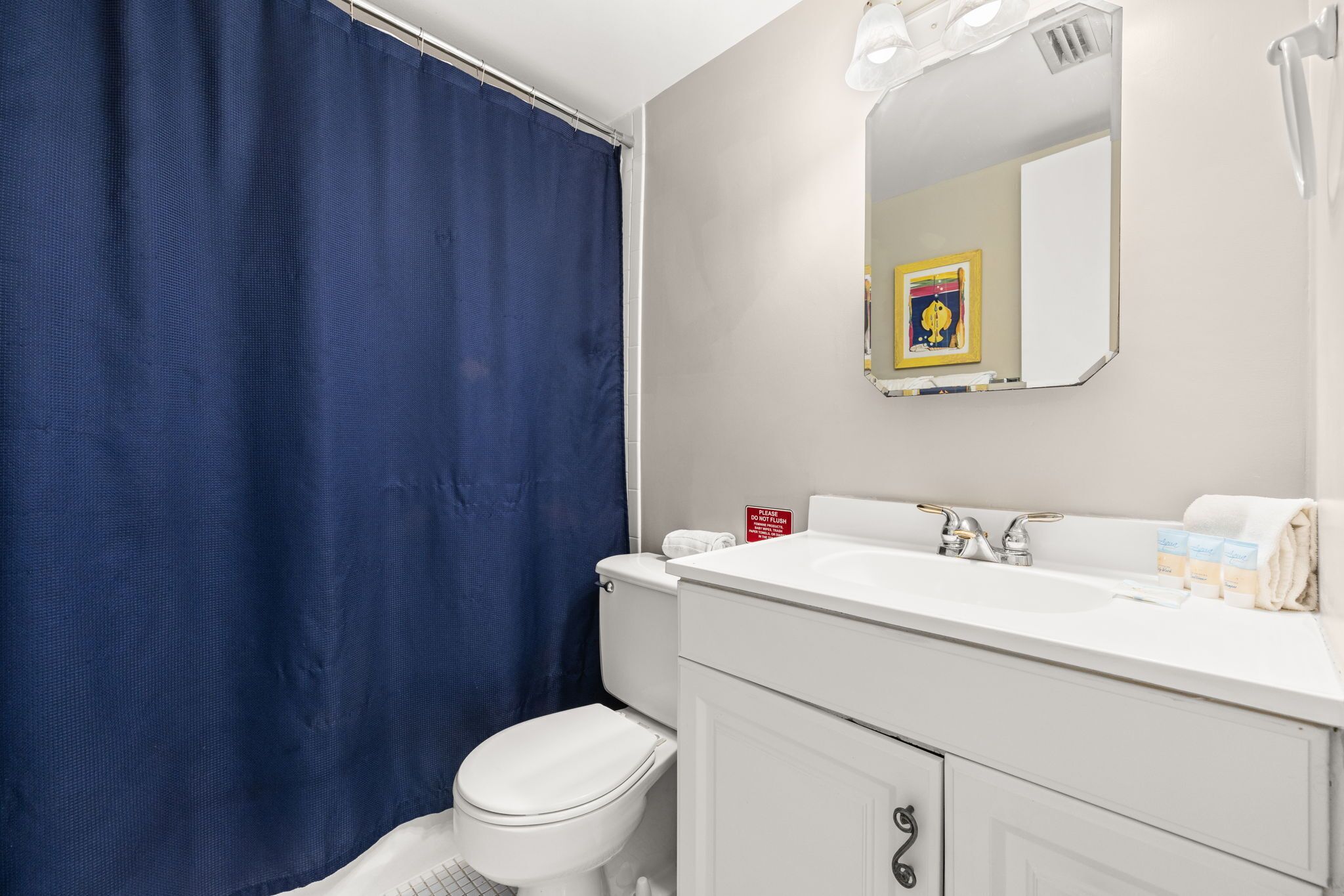 A bathroom with a white toilet, a blue shower curtain, a white sink with a mirror above it, and a sign on the wall.
