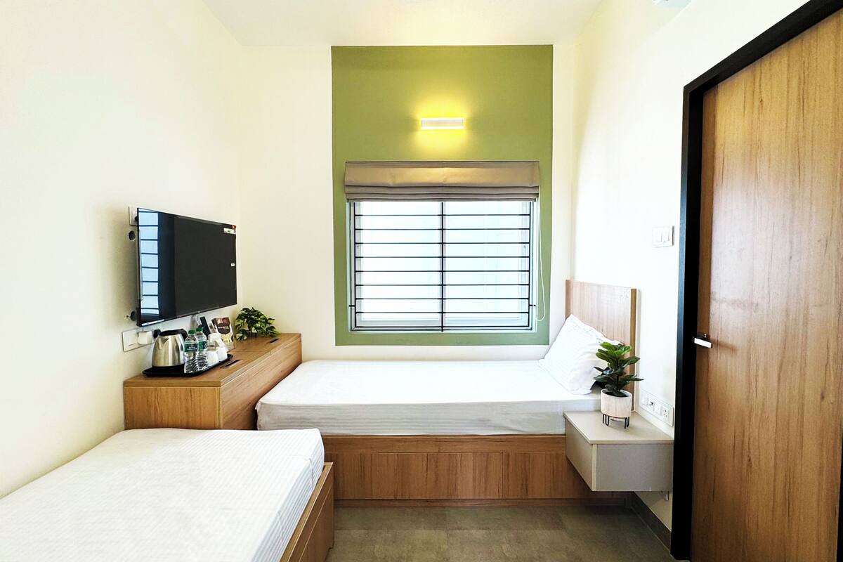 Habitación de hotel pequeña y funcional con una cama individual, una mesita de noche con una planta, un televisor montado en la pared y una ventana con persianas.
