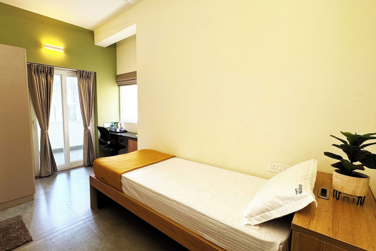 Una habitación pequeña con una cama individual, un escritorio, una silla, un ventanal con cortinas y una planta en maceta.