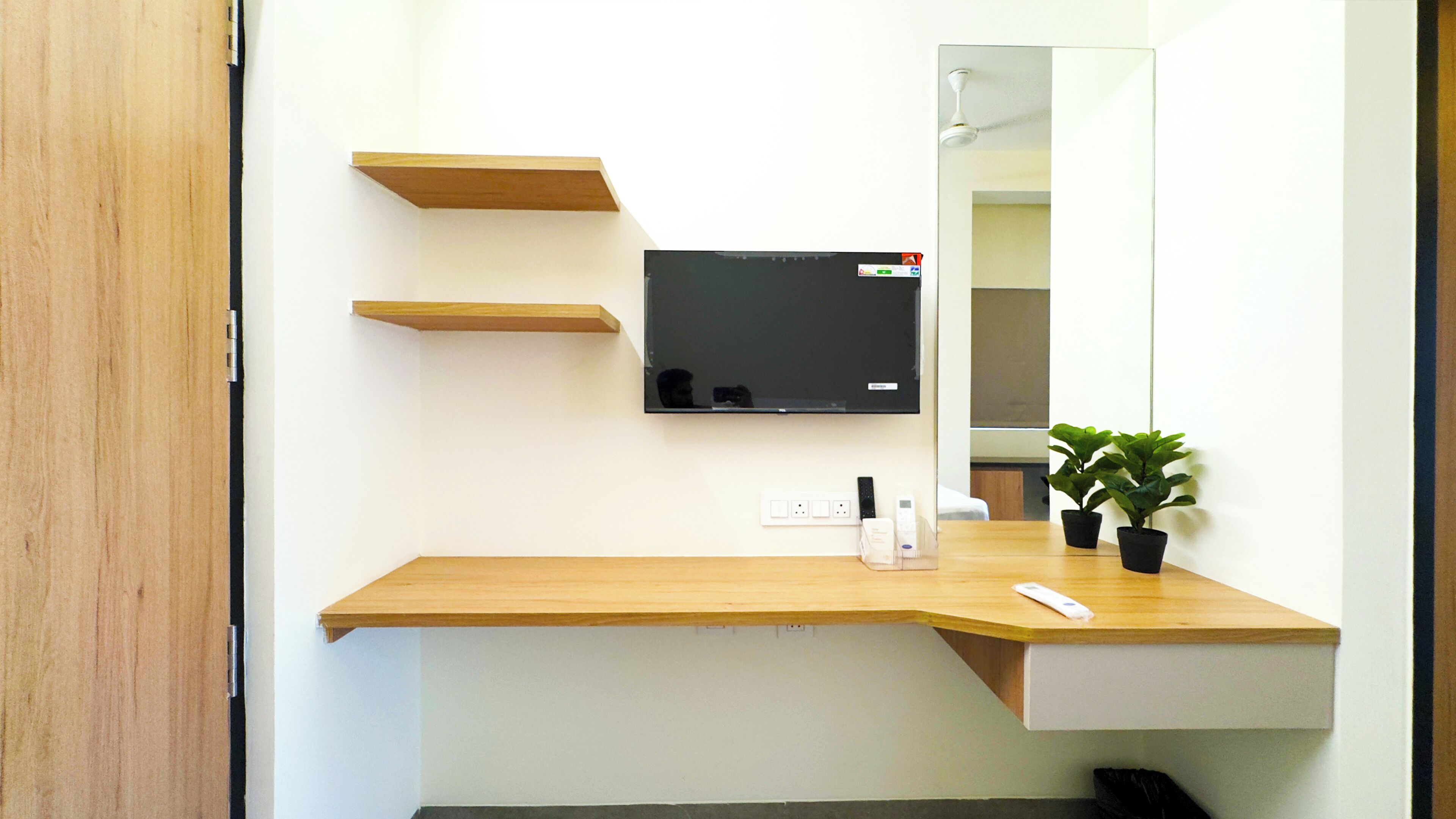 A modern bathroom with a wooden vanity, a mounted flat-screen TV, a mirror, and two potted plants.
