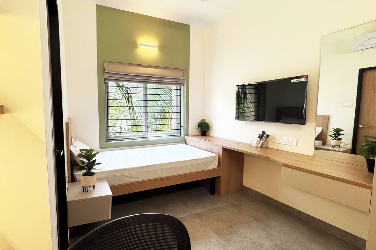 Una habitación pequeña y moderna con escritorio, una banca, televisor y ventana con vista a árboles.