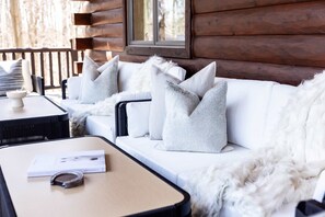 Ein rustikal eingerichtetes Blockhauszimmer mit einer weißen Couch und weichen, weißen Zierkissen, einem kleinen Tisch mit einem Buch und einer Kamera sowie einem Fenster mit Blick auf Bäume.