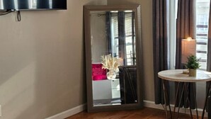 Et værelse med trægulv, et fladskærms-tv ophængt på væggen, et lille rundt bord med et stearinlys og et spejl der afspejler en pink stol og en plante.