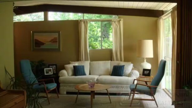 A living room with a sofa, two armchairs, a coffee table, and a lamp. There is a window with curtains and a painting on the wall.