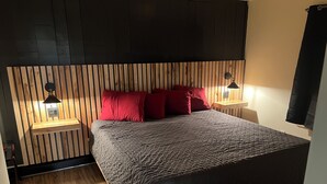 A bedroom with a wooden slat headboard, a bed with a textured grey bedspread, red pillows, and two bedside lamps on wooden nightstands. - Comfort and warmth, convenience to Campus at Pike Bay Apartment (Chassell)