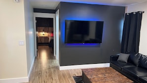A modern living room with a flat-screen TV mounted on a dark wall, a black leather sofa, and a wooden coffee table. - Comfort and warmth, convenience to Campus at Pike Bay Apartment (Chassell)