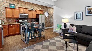 Una cocina moderna con armarios de madera, microondas y refrigerador. Hay un comedor con una mesa y sillas. Un sofá negro y una pequeña mesa de centro están en la sala.