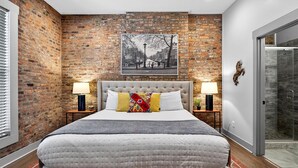 Un dormitorio con pared de ladrillo, una cama grande con ropa blanca, dos mesitas de noche con lámparas, y un cuadro colgado sobre la cama.