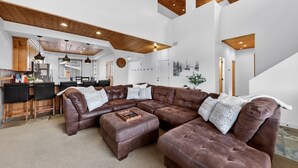 Un'ampia zona giorno con un grande divano in pelle marrone a forma di L, un soffitto in legno e una cucina sullo sfondo.