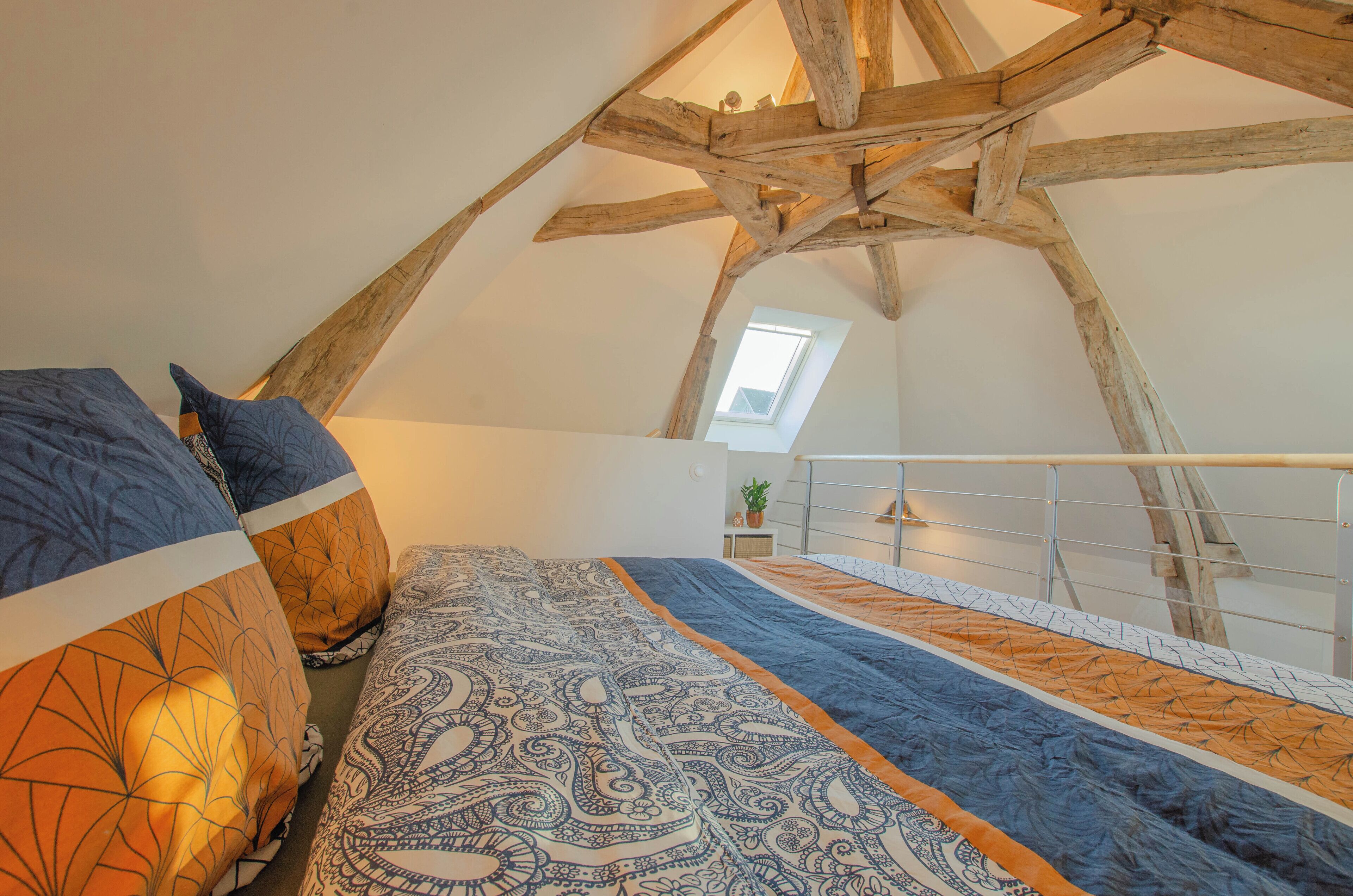 Ein Schlafzimmer mit schrägen Wänden, einem Oberlicht, einem Bett mit gemusterten Betttextilien und einer kleinen Pflanze auf einem Nachttisch.