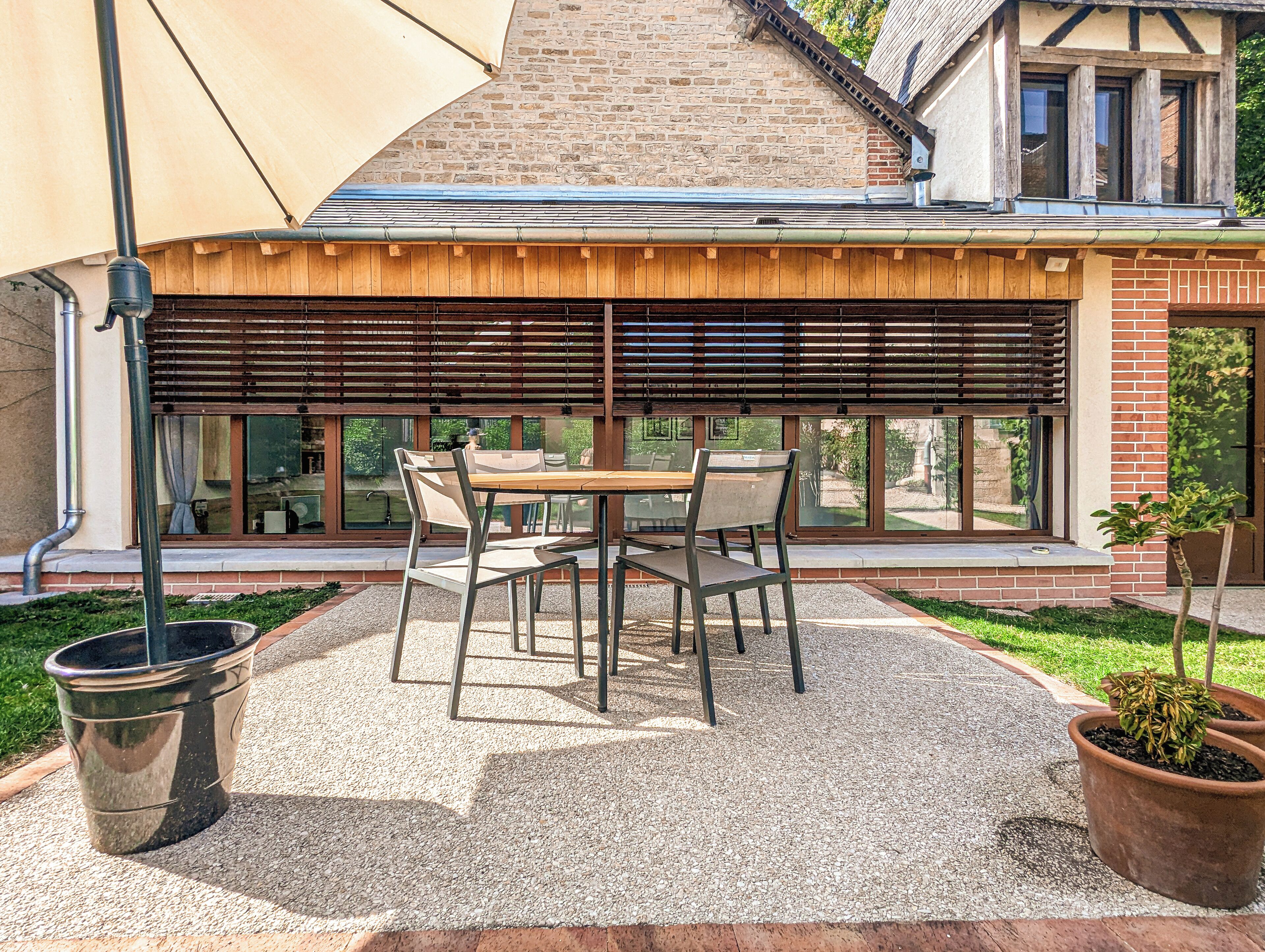 Une terrasse avec une table et des chaises, un pot de fleurs et un bâtiment doté de grandes fenêtres et d’une façade en briques.