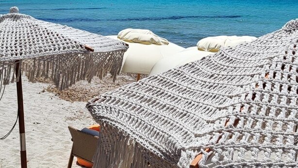 Una playa con sombrillas de macramé y sillas de descanso, una mesa de madera y vistas al mar y montañas distantes.