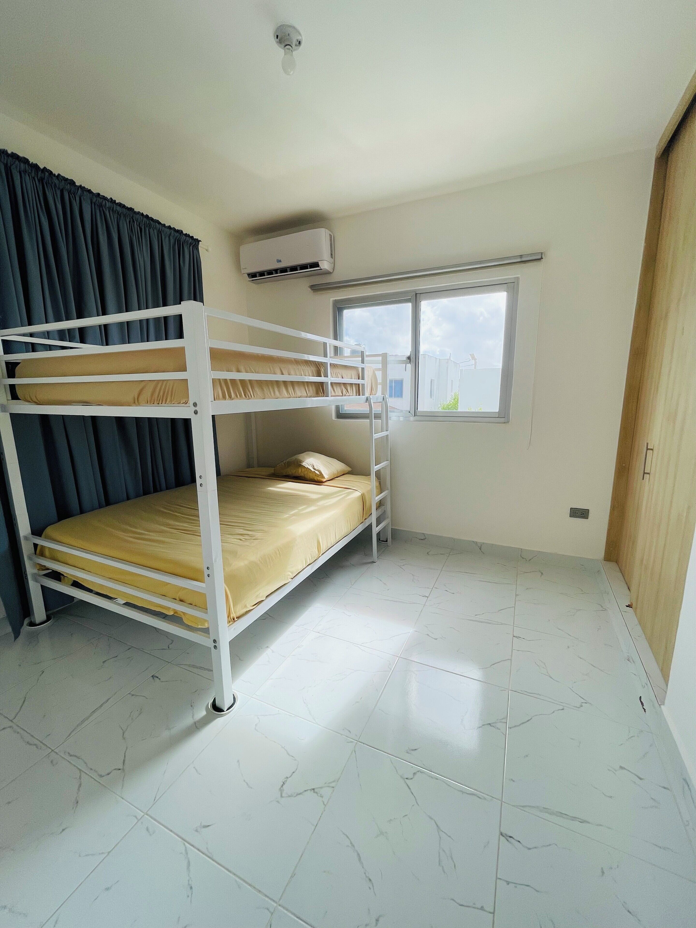 Een slaapkamer met stapelbed, raam, airconditioning en betegelde vloer.