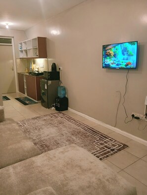 A living room with a sofa, a coffee table, a TV mounted on the wall, a kitchen area with cabinets and a refrigerator, and a patterned rug on the floor. - Fantastic Executive Studio apartment in welcoming Nairobi for your stay (Nairobi)