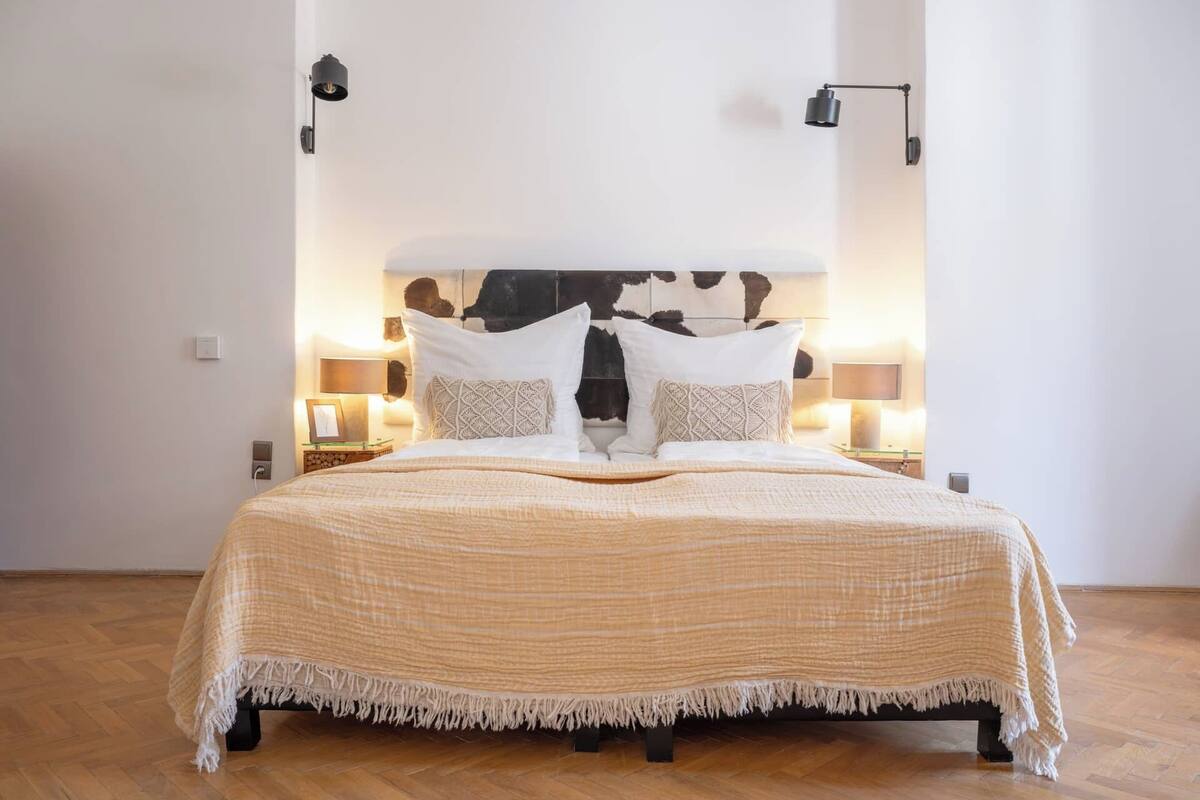 A neatly made bed with a beige blanket and pillows, two bedside lamps, and a wooden floor.