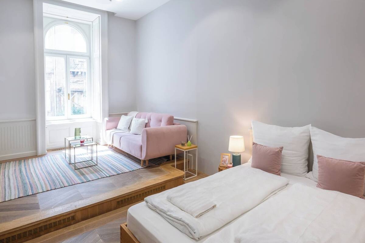 A bedroom with a bed, a sofa, a nightstand, a lamp, and a window with a view of buildings.