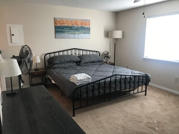 A bedroom with a bed, bedside tables, a lamp, a fan, and a painting on the wall.