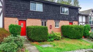 Un immeuble résidentiel à deux étages, doté d’une porte rouge, d’une façade en briques et d’un jardin bien entretenu avec des haies.