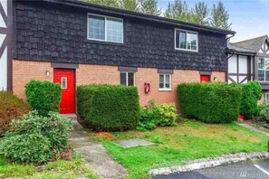 A two-story residential building with a red door, a brick facade, and a well-maintained front yard with hedges.