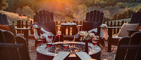 A circular outdoor seating area with Adirondack chairs and a fire pit, surrounded by a fence and a view of a hilly landscape.