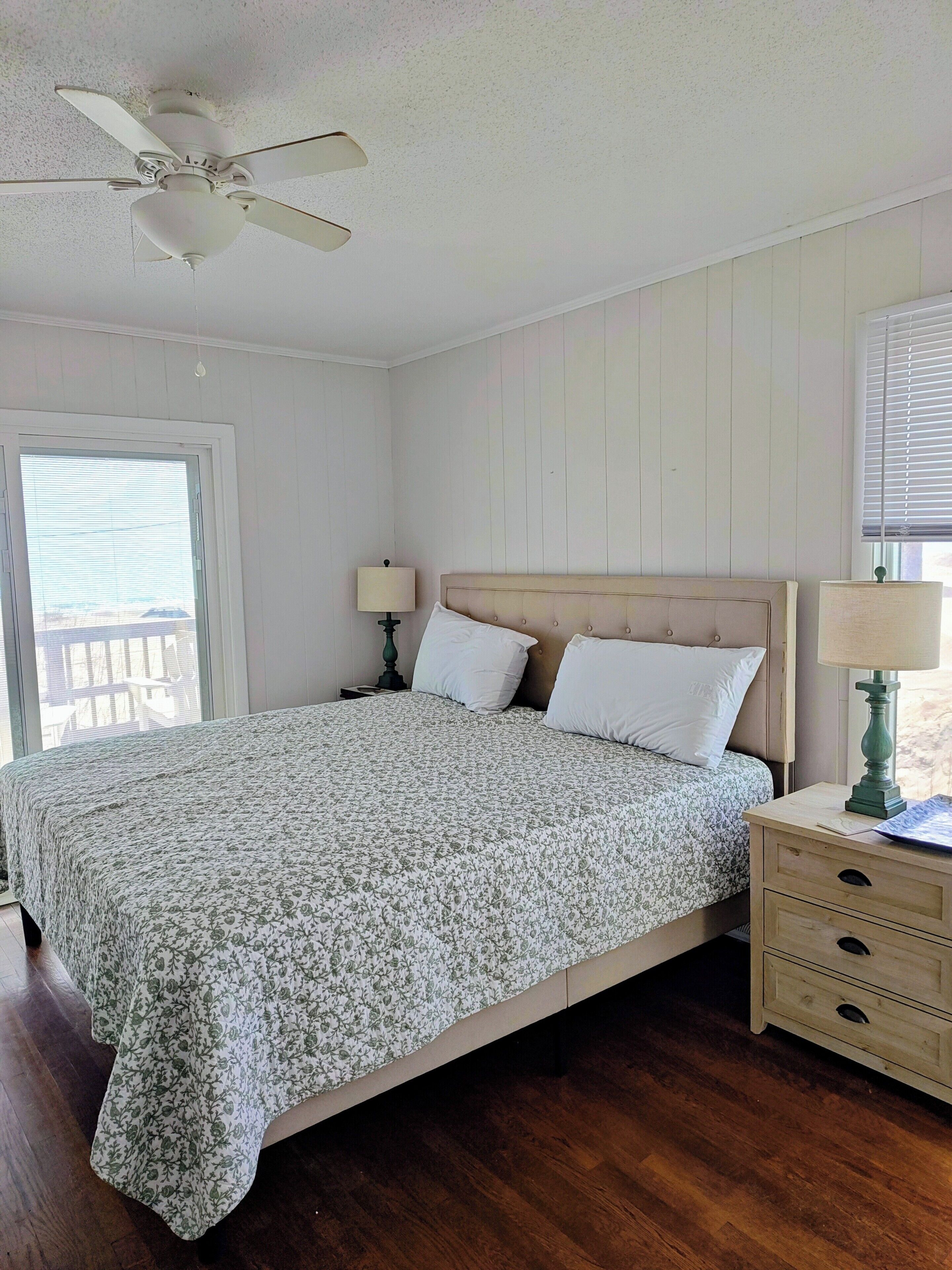 A bedroom with a bed, a nightstand, a lamp, a ceiling fan, and a window with blinds.