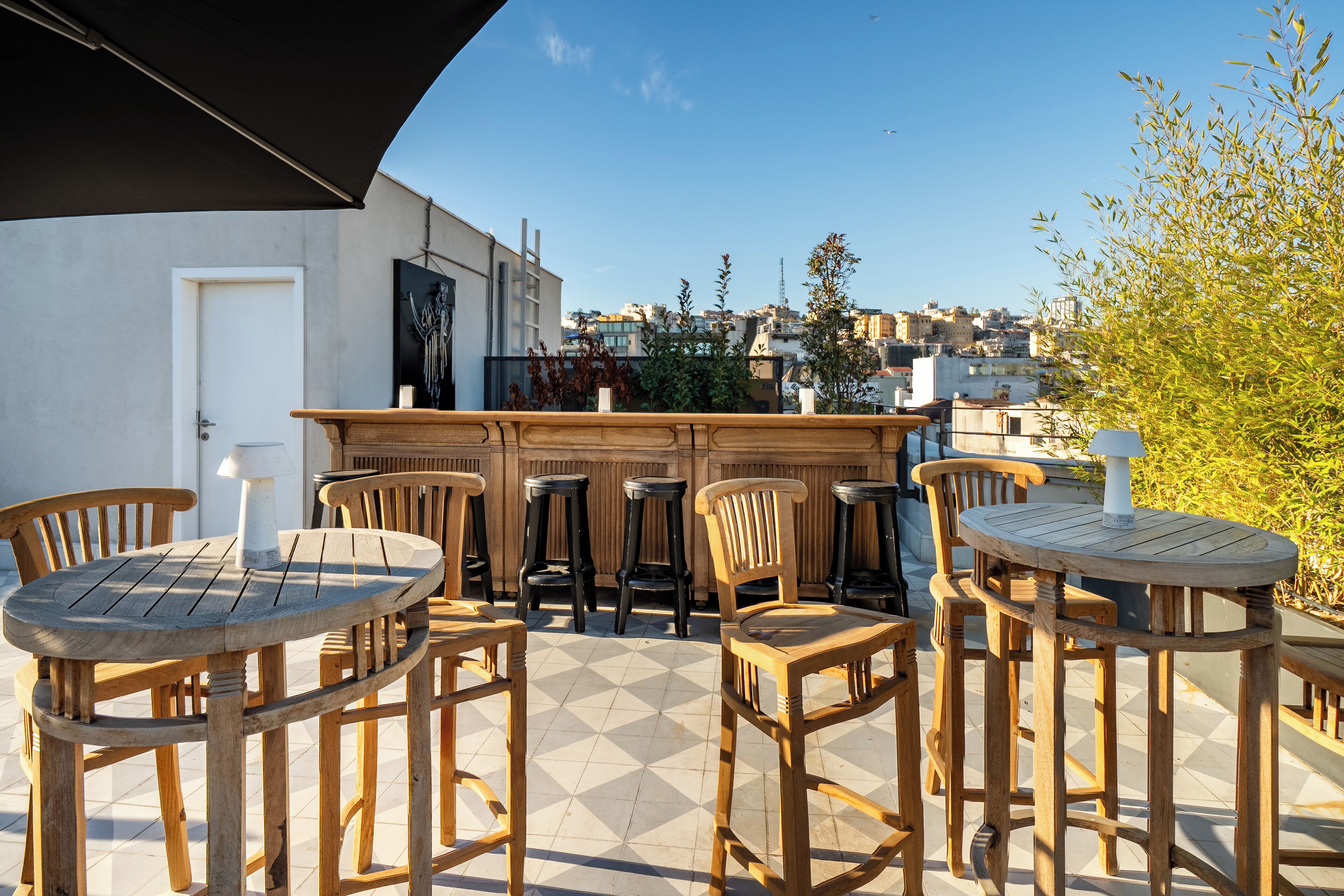 Een terras buiten met houten tafels en stoelen, een bar en een uitzicht op de stad.