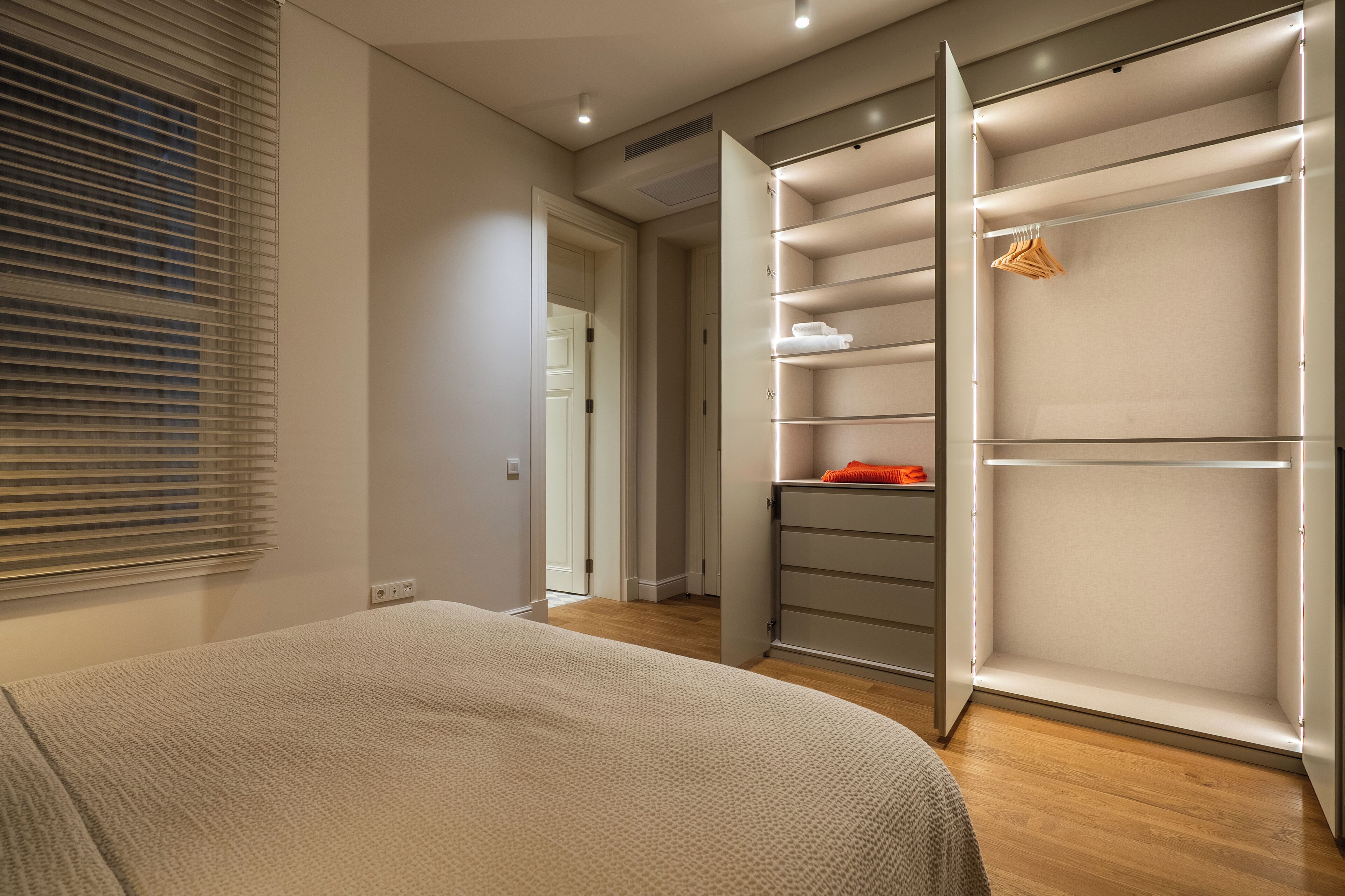 Une chambre à coucher avec un lit, un dressing avec des étagères et une fenêtre avec des stores.
