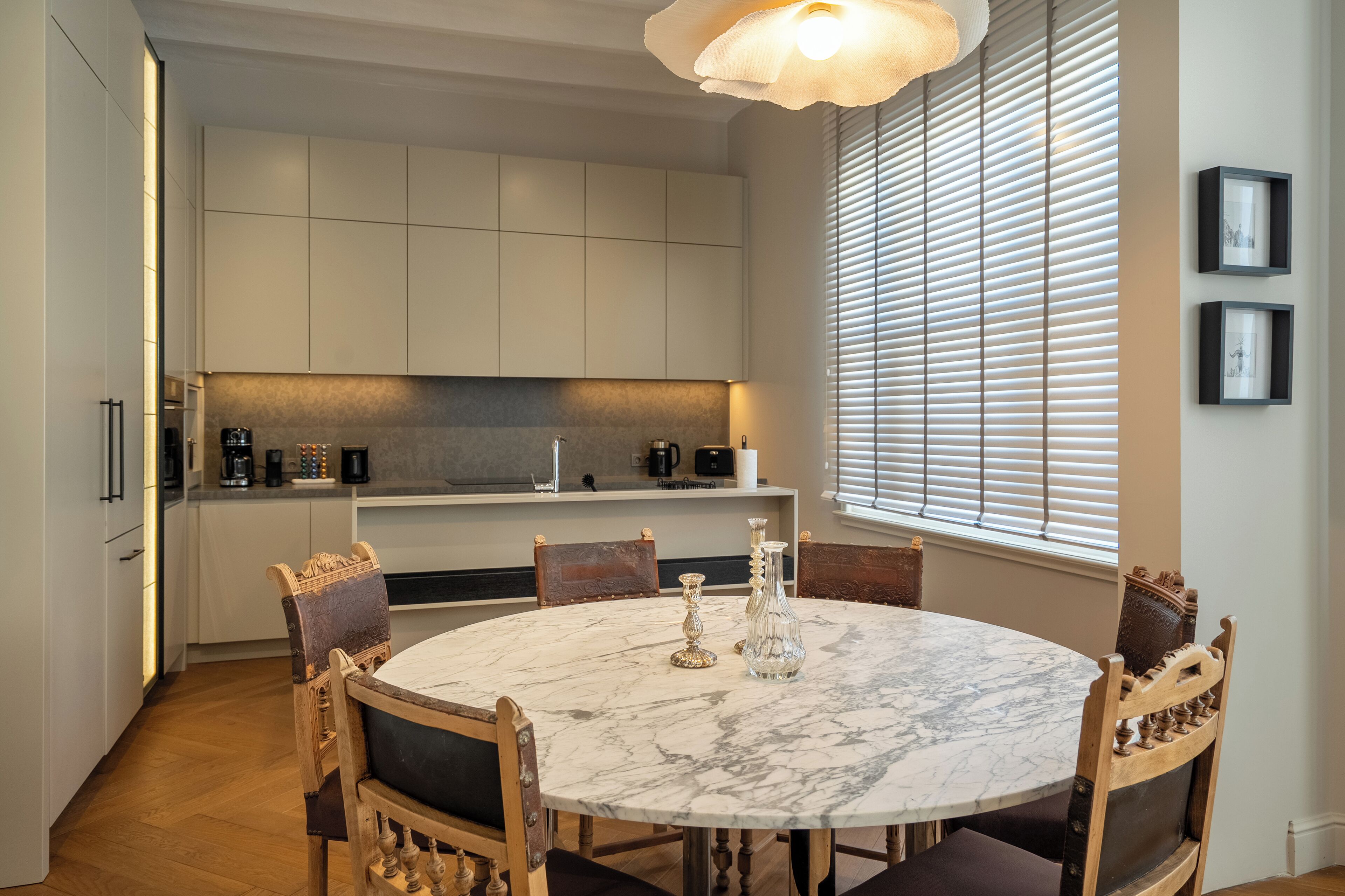 Een moderne keuken met witte kasten, een marmeren eettafel en een groot raam met zonwering.