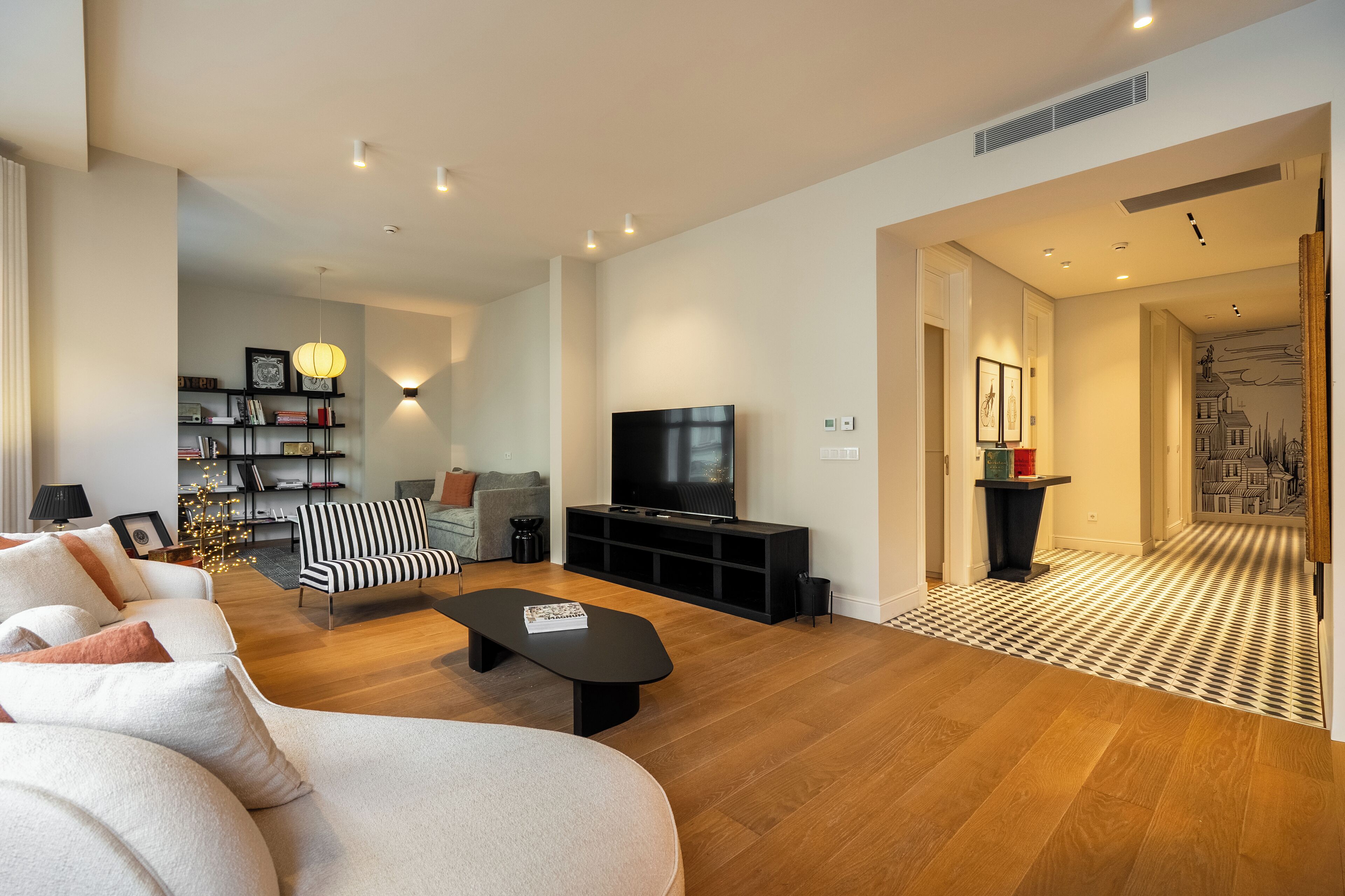 Una sala de estar moderna con un sofá, una mesa de centro negra, un sillón a rayas, un televisor sobre un mueble negro y una estantería.