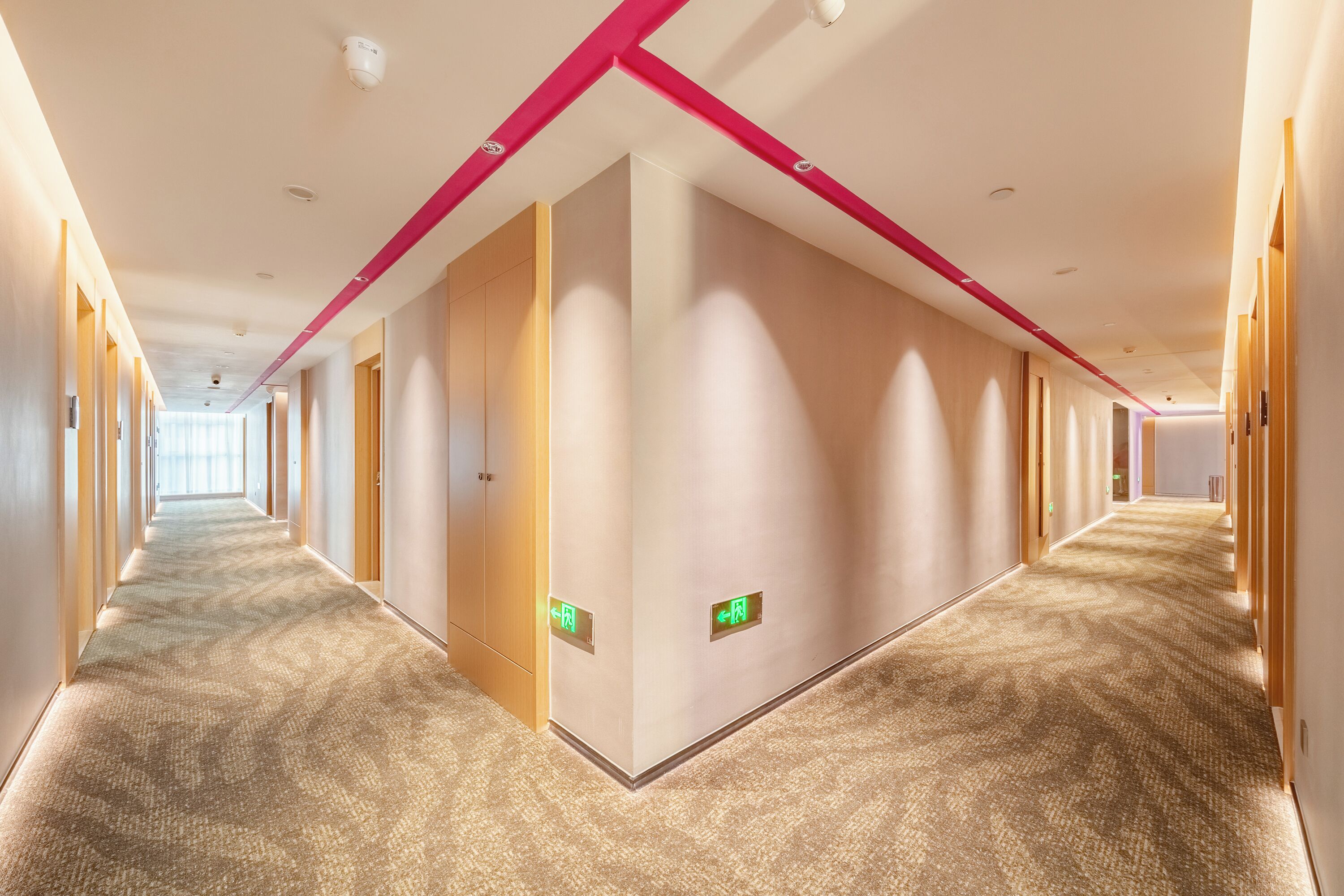 A long, narrow hotel corridor with carpeted floors, wooden doors, and a pink ceiling beam.