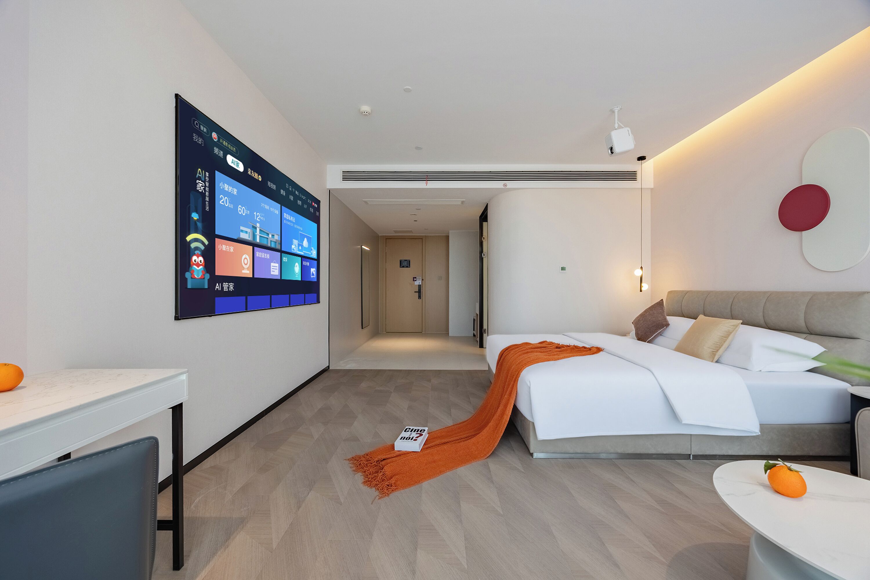 Een moderne hotelkamer met een groot scherm aan de muur, een bed met een witte dekbedovertrek en een oranje plaid, een tafeltje met een stuk oranje fruit, en een bank met een kussen.
