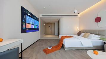 Een moderne hotelkamer met een groot scherm aan de muur, een bed met een witte dekbedovertrek en een oranje plaid, een tafeltje met een stuk oranje fruit, en een bank met een kussen.