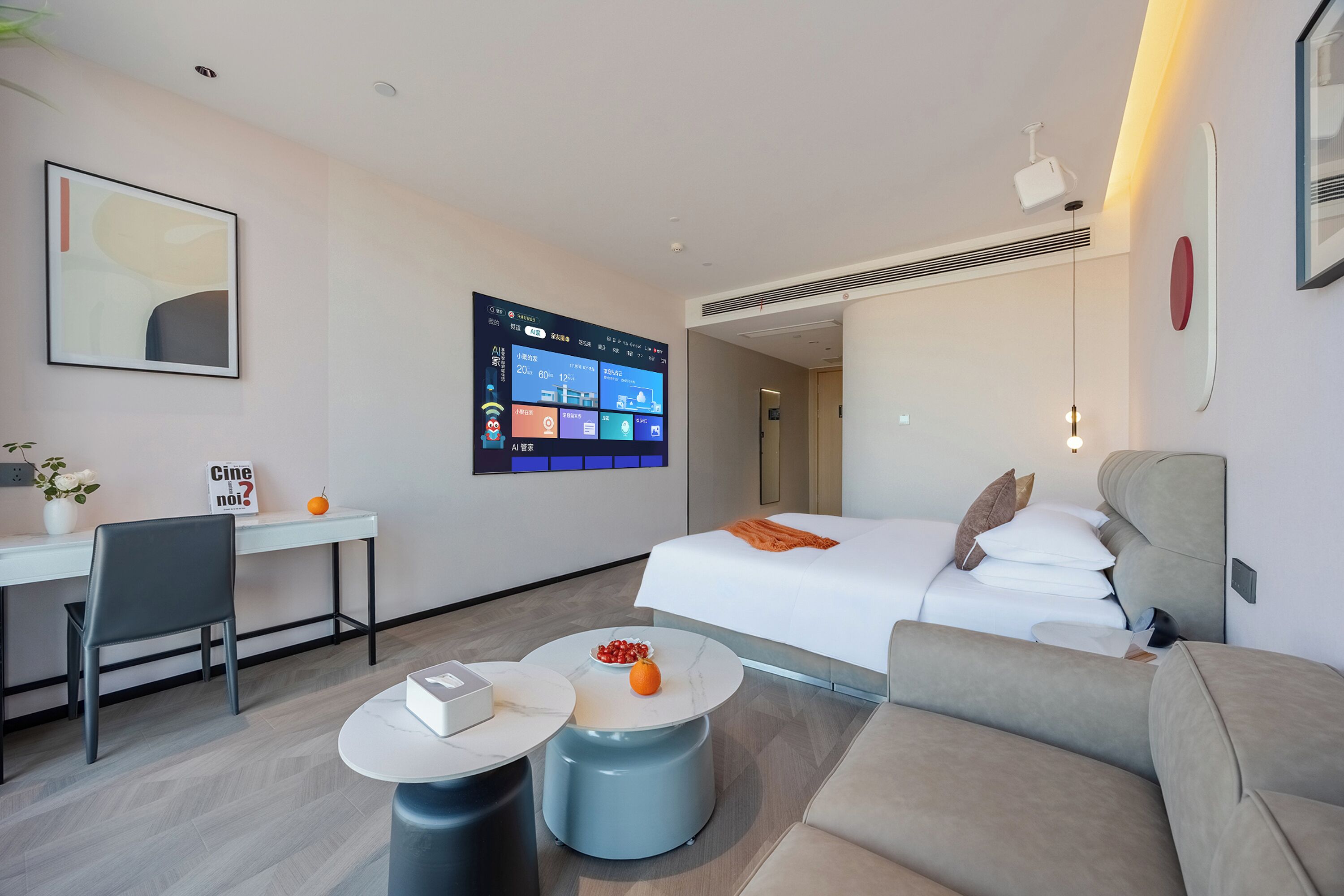 A modern hotel room with a bed, a desk, a chair, a coffee table, and a large digital display on the wall.