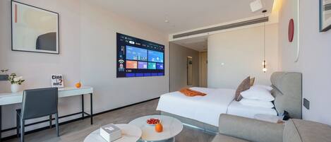A modern hotel room with a bed, a desk, a chair, a coffee table, and a large digital display on the wall.
