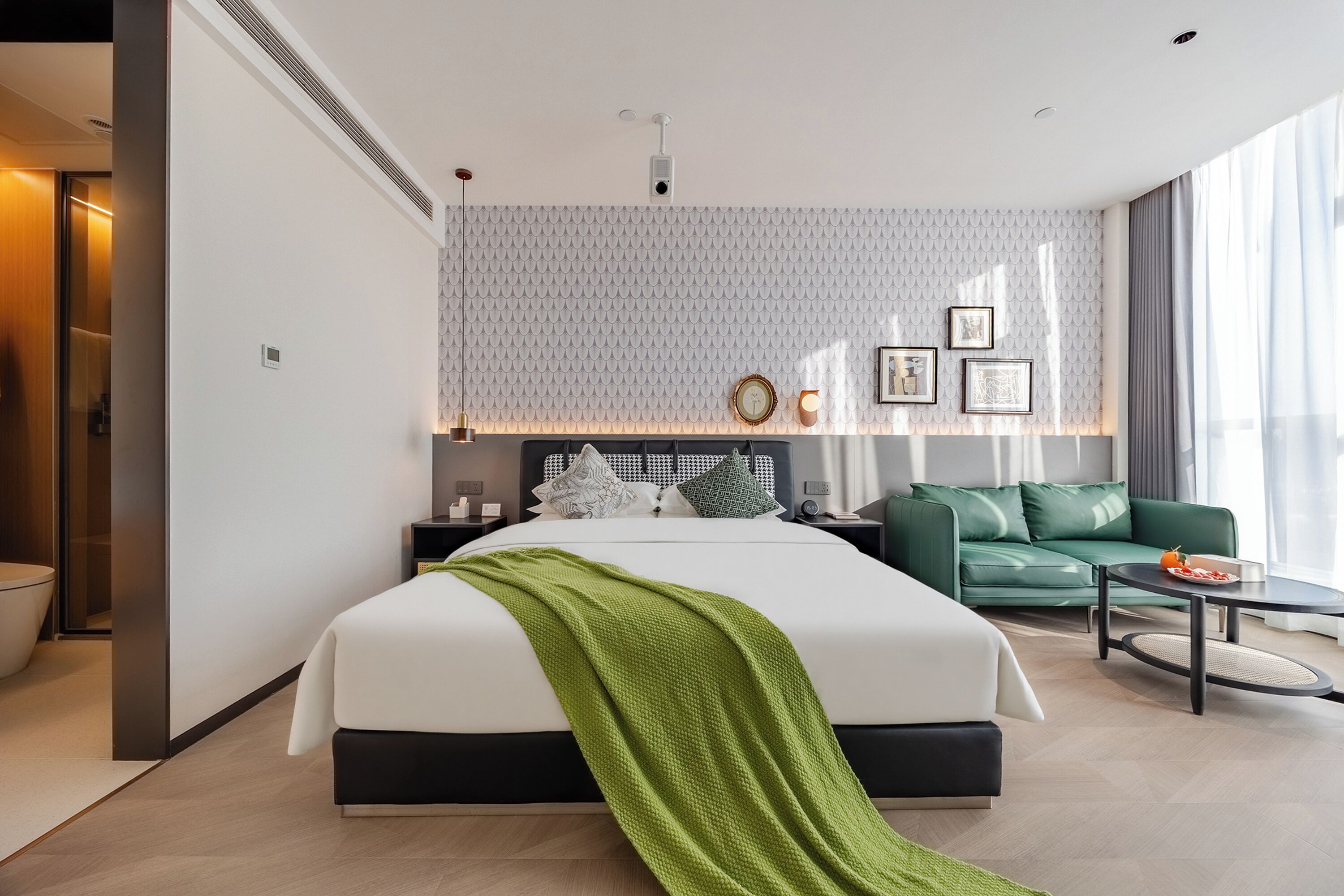 A modern hotel room with a large bed, a green sofa, a round coffee table, and a patterned wall.