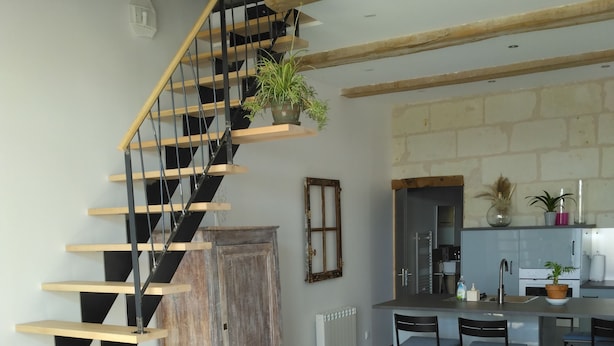 Un salon moderne avec un canapé d’angle gris, un escalier en bois et un coin cuisine doté d’un comptoir et de chaises.