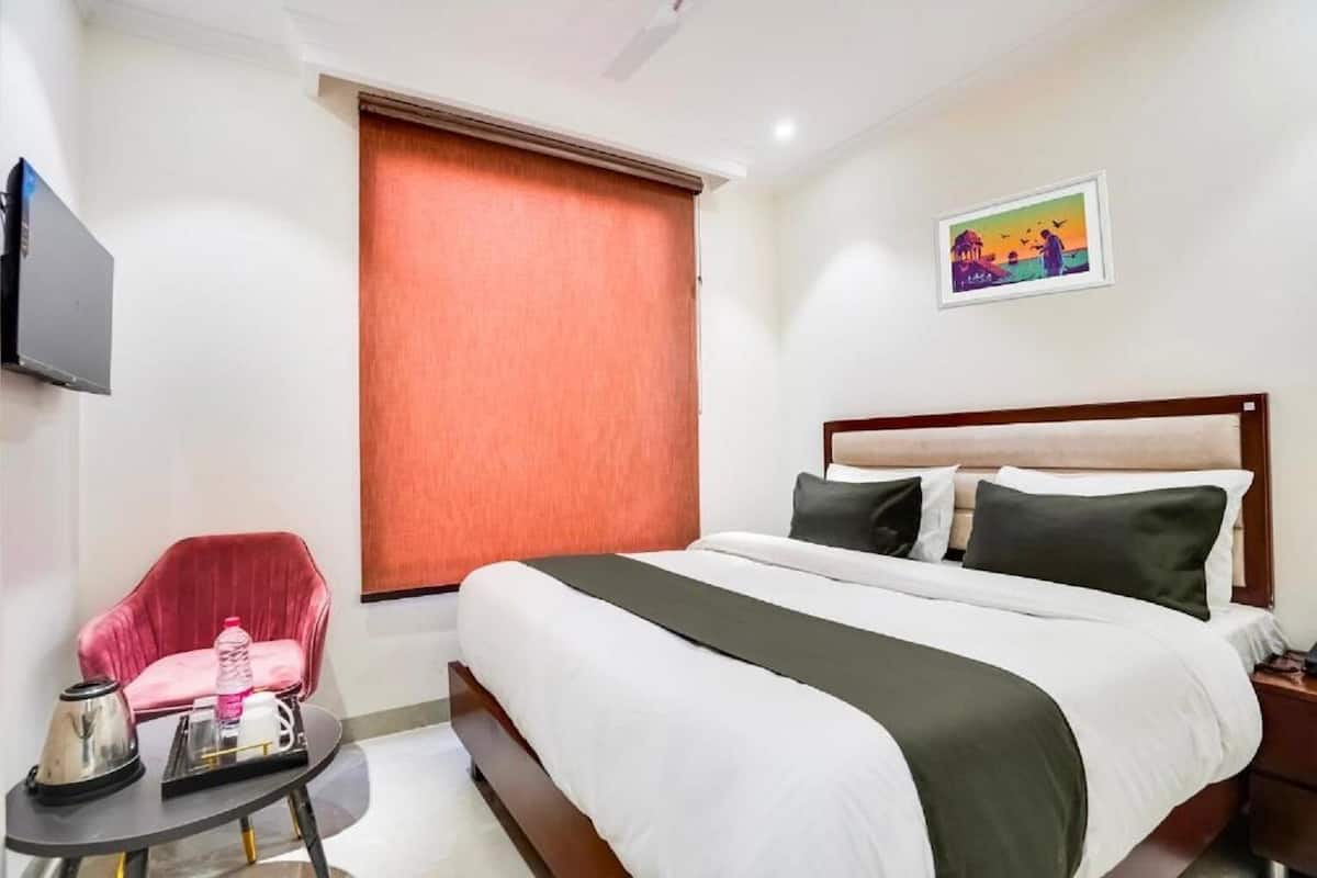 Habitación de hotel con cama, una silla, una mesita con hervidor y botella, un cuadro en la pared y una ventana con persianas.