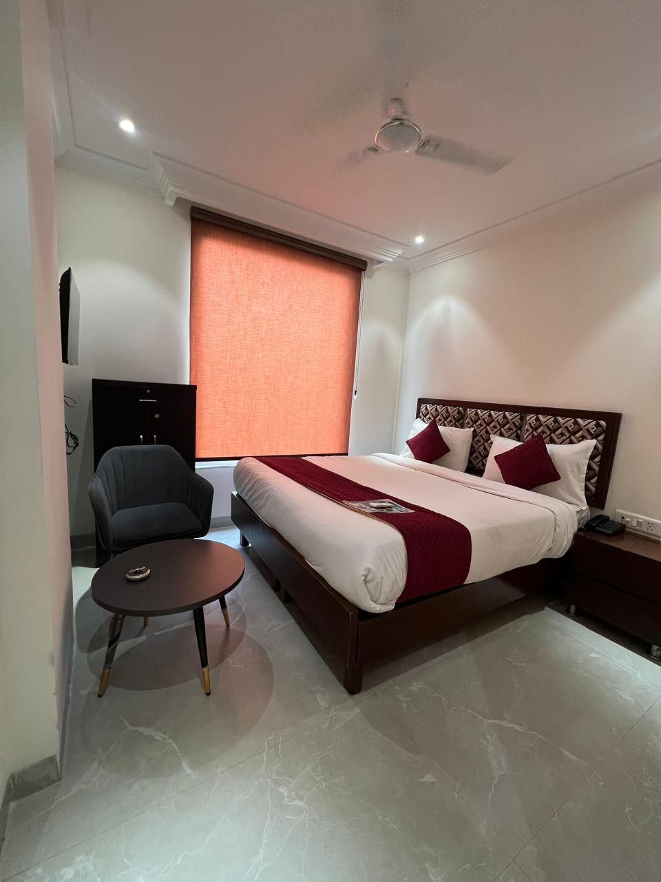Habitación de hotel con cama, una silla, una mesita, ventilador de techo y ventana con persianas.