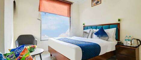 A hotel room with a bed, a chair, a nightstand, a window with a view, and a small table with snacks.
